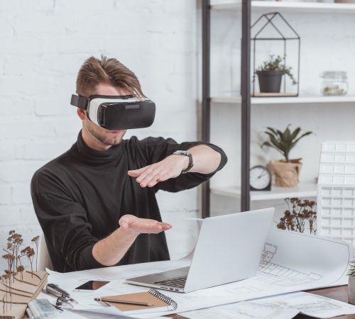 architect in vr headset at workplace with laptop, schemes and building models in office